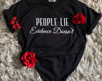 May include: Black t-shirt with the text "PEOPLE LIE Evidence Doesn't" in white. The shirt has a crew neck and short sleeves. Red roses are placed around the shirt. The brand "Before the Crypt" is printed on the collar.