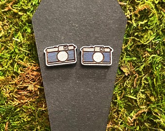 May include: A pair of wooden camera earrings. The earrings are painted blue with white details. The earrings are displayed on a black coffin-shaped display card.