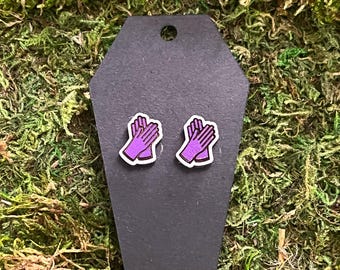 May include: A pair of purple glove earrings with a white outline, displayed on a black hexagonal card. The earrings are set against a backdrop of green moss.