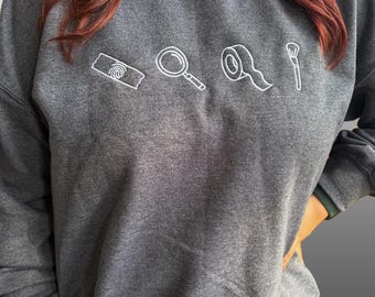 May include: A dark gray sweatshirt with white embroidered illustrations of a roll of tape, a magnifying glass, a roll of tape, and a makeup brush. The sweatshirt is a crew neck style.