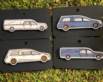 May include: Four wooden laser-cut pins of hearses in different colors. The hearses are white, blue, white, and blue. The pins are displayed on black card stock.