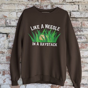 Może przedstawiać: Brązowa bluza z nadrukiem "Like a needle in a haystack" na przodzie. Tekst jest biały i otoczony zieloną trawą.