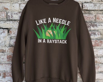 May include: A brown sweatshirt with the text "Like a needle in a haystack" printed on the front. The text is in white and is surrounded by green grass.
