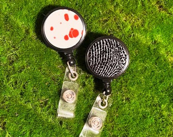 May include: Two black retractable badge holders with clear straps. One holder has a white background with red splatter paint. The other holder has a black and white geometric pattern.