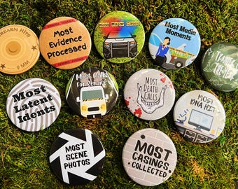 May include: Ten round buttons with different designs and text. The buttons are white, black, green, and beige with black text. The text on the buttons includes "Most Firearm Hits", "Most Evidence Processed", "Most Major Cases", "Most Media Moments", "Most Latent Idents", "Most Calls for Service", "Most Death Calls", "Trainer of the Year", "Most DNA Hits", "Most Scene Photos", and "Most Casings Collected".