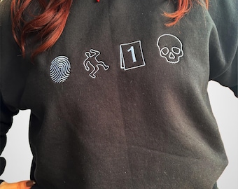 May include: Black sweatshirt with white embroidered crime scene icons: a fingerprint, a body outline, a numbered evidence marker, and a skull. The sweatshirt is a crew neck style.