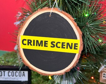 May include: A round wooden Christmas ornament with a black background and a yellow "CRIME SCENE" tape across the middle.