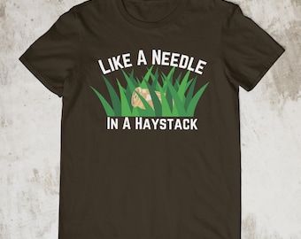 May include: A brown t-shirt with the text "Like a needle in a haystack" printed in white. A small illustration of a needle in a patch of green grass is above the text.