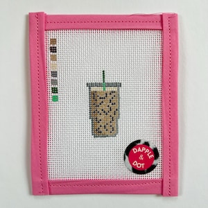 May include: Needlepoint canvas featuring a coffee drink design. The canvas is framed in a bright pink fabric. A color palette of brown, tan, and green is visible on the left side. A sticker with the text "Dapple & Dot" is on the bottom right.