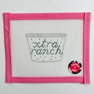 May include: A needlepoint canvas with a pink border and the words "extra ranch" stitched in green. The design features a gray container, and a small round sticker with the words "Dapple & Dot".