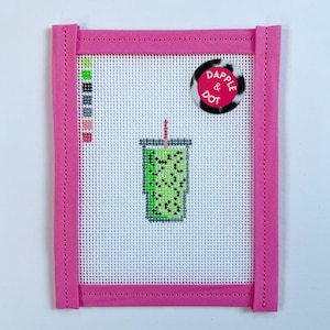 May include: A needlepoint canvas featuring a green drink with black speckles and a red straw. The canvas is framed in bright pink fabric. A circular sticker with the text "Dapple & Dot" is in the upper right corner.