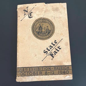 May include: Vintage State Fair program from Raleigh, North Carolina, featuring the state seal and the text "State Fair." The cover is aged with visible wear and tear, and the date "October 8-12 1940" is printed at the bottom.