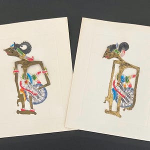 May include: Two framed Indonesian shadow puppets. The puppets are cut from a gold-colored material and feature detailed painted designs in red, blue, green, and white. The puppets are mounted on white paper.