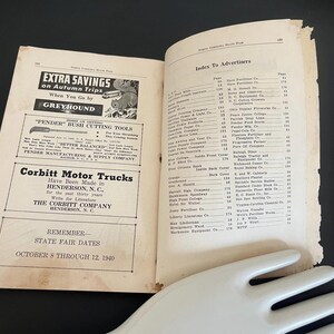 May include: Open vintage book with advertisements, including Greyhound bus travel and Pender Bush Cutting Tools. The right page lists an index of advertisers. A white, hand-shaped serving piece rests at the bottom.