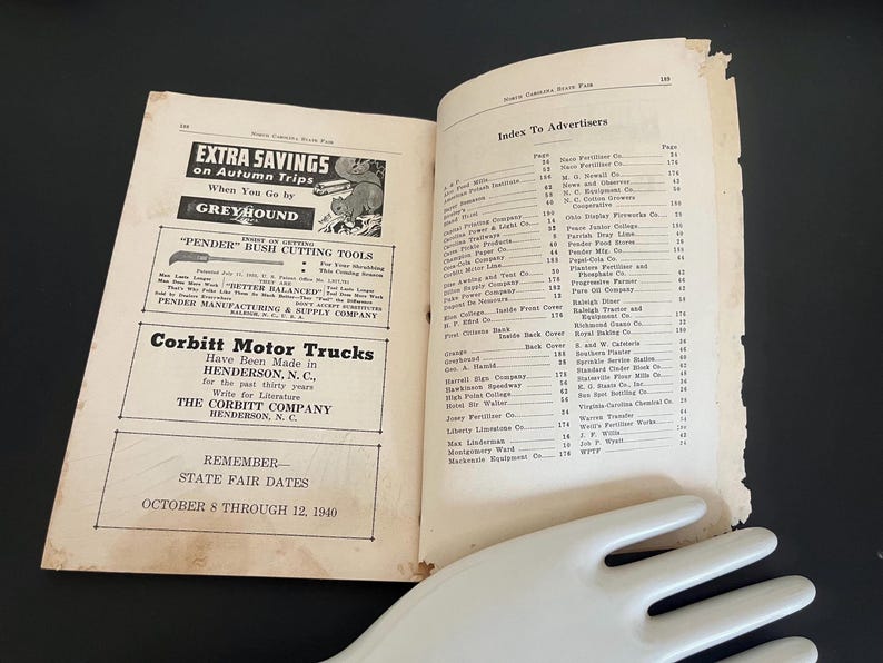 May include: Open vintage book with advertisements, including Greyhound bus travel and Pender Bush Cutting Tools. The right page lists an index of advertisers. A white, hand-shaped serving piece rests at the bottom.