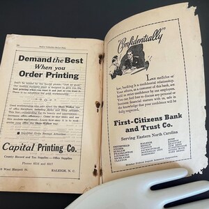 May include: An open vintage book with aged, yellowed pages. The left page features text about ordering printing, while the right page advertises First-Citizens Bank and Trust Co. The book is open on a dark surface.