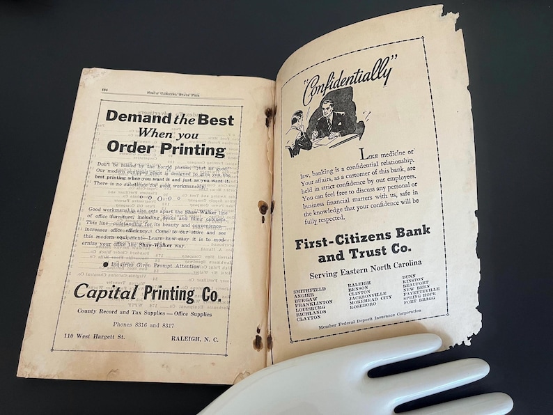 May include: An open vintage book with aged, yellowed pages. The left page features text about ordering printing, while the right page advertises First-Citizens Bank and Trust Co. The book is open on a dark surface.