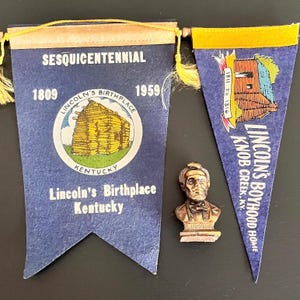 May include: A collection of historical memorabilia featuring Abraham Lincoln. Includes two blue felt pennants with gold trim and text about Lincoln's birthplace and boyhood home in Kentucky, plus a bronze bust of Lincoln.