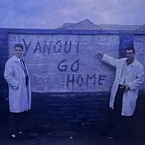 May include: Two individuals wearing light-colored trench coats stand on either side of a stone wall. The wall features the hand-painted text YANQUI GO HOME in dark, uneven lettering. The scene is captured in a cool, blue-toned photographic style.