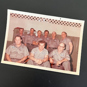 May include: A group of eight men wearing gray work shirts with a logo on the left chest. The men are standing in a locker room with lockers in the background. Some of the men are smiling.