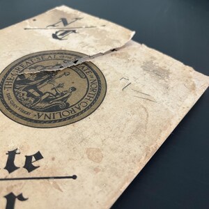 May include: A vintage document with the words "THE GREAT SEAL OF THE STATE OF NORTH CAROLINA" and the state seal. The document is tan with dark brown text and a black anchor symbol. The paper is torn and shows signs of age.