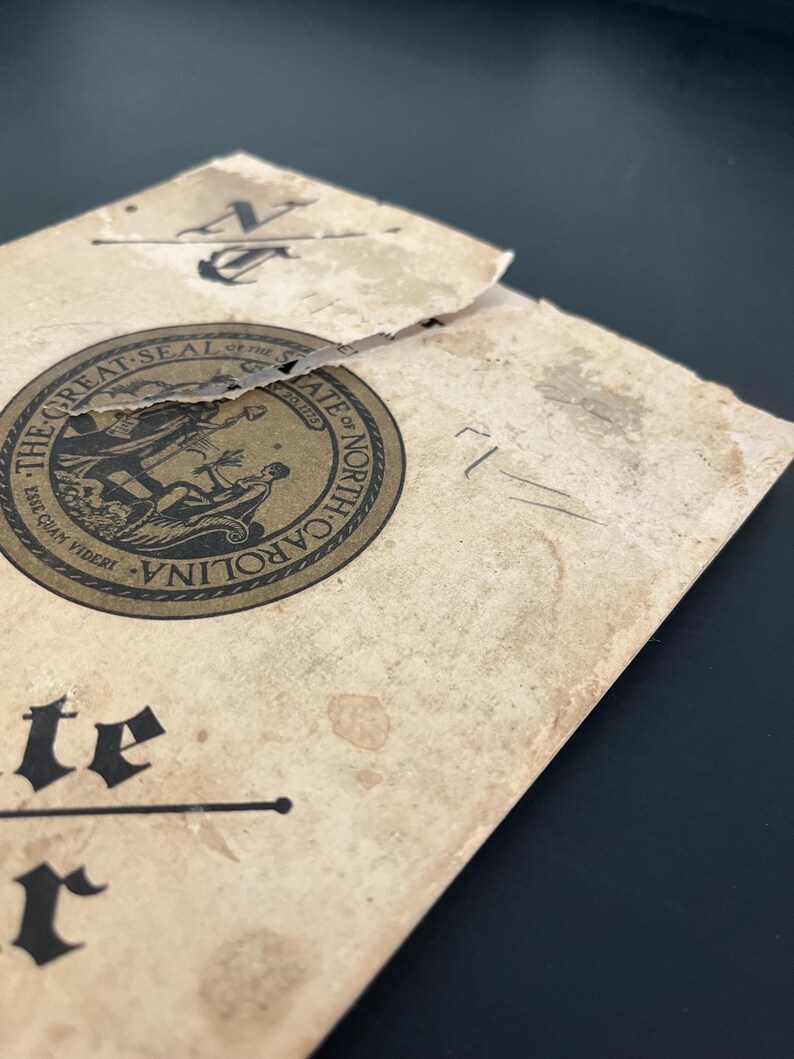 May include: A vintage document with the words "THE GREAT SEAL OF THE STATE OF NORTH CAROLINA" and the state seal. The document is tan with dark brown text and a black anchor symbol. The paper is torn and shows signs of age.