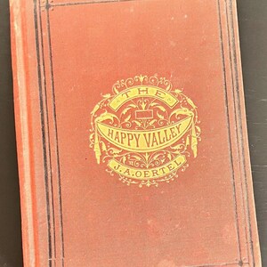 May include: A vintage book titled "The Happy Valley" by J.A. Oertel. The cover is a muted reddish-orange with a decorative gold emblem and black border. The book's spine is visible on the left side.