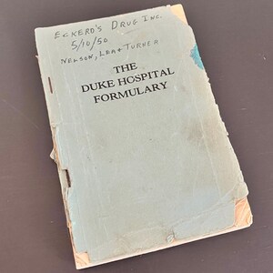 May include: A vintage book titled "The Duke Hospital Formulary" with the text "Eckerd's Drug Inc." and the date "5/10/50" handwritten on the cover. The book has a light blue cover with visible wear and tear.
