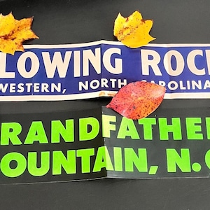 May include: Two rectangular souvenir bumper stickers. The top sticker is blue with white text reading BLOWING ROCK WESTERN, NORTH CAROLINA. The bottom sticker is black with bright green text reading GRANDFATHER MOUNTAIN, N.C. Four autumn leaves are placed around them.