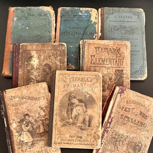 May include: A collection of eight antique hardcover schoolbooks with worn, distressed covers in shades of blue, brown, and tan. The books feature vintage typography and illustrations, including titles like The Elementary Spelling-Book and Venable's Primary Arithmetic.