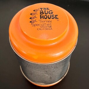 May include: An orange container with a mesh body and a matching lid. The lid features the text "THE BUG HOUSE" and "Tierney Specialties STOCKTON CALIFORNIA". The container is cylindrical and appears to be made of metal and plastic.
