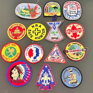 May include: A collection of vintage patches with various designs and text. Patches include images of a deer, a bear, a Native American, and text such as "Camporee", "NRA Safe Hunter", and "Boy Power". The patches are in various shapes and colors.