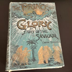 May include: Antique book titled "The Prince of Glory, Story of the Saviour" by Canon Farrar. The cover features gold and teal illustrations of religious figures, palm trees, and text. The book has a worn, vintage appearance.