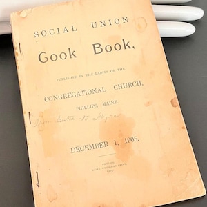 May include: A vintage softcover booklet titled Social Union Gook Book, published by the Ladies of the Congregational Church in Phillips, Maine, dated December 1, 1905. The cover is light tan with dark text and visible age-related staining.