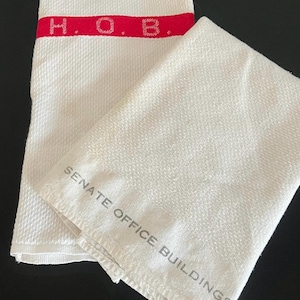 May include: Two white towels. One towel has a red stripe with the letters "H.O.B." The other towel has the words "SENATE OFFICE BUILDINGS" printed on it. Both towels are folded and have a textured weave.