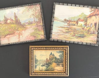 May include: Three framed vintage landscape paintings with black, gold, and white frames. The paintings depict a river scene, a village scene, and a castle scene.