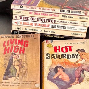 May include: A stack of vintage paperback books with colorful covers. Titles include "Tarzan and the Leopard Men," "The Green Wound Contract," and "Hot Saturday." The books are stacked on a dark surface, showcasing their aged appearance.
