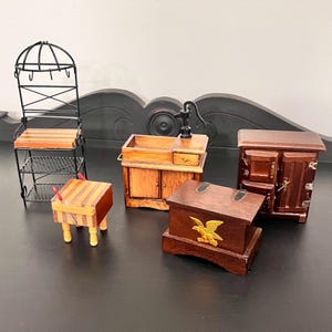 May include: A collection of miniature wooden furniture pieces, including a shelf, a table, a cabinet, a chest with a gold eagle emblem, and a sink with a pump. The pieces are in various shades of brown and are arranged on a dark surface.