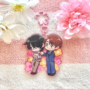 May include: A clear acrylic keychain featuring two anime-style characters standing together. One character has black hair and wears a tan suit, while the other has brown hair and wears a blue pinstripe suit. The background includes pink and yellow flowers.