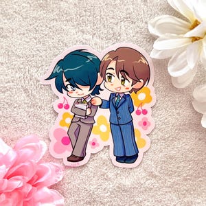 Op de afbeelding: Een sticker met twee anime-achtige personages in pakken. Eén heeft turquoise haar en houdt papieren vast, de ander heeft bruin haar en draagt een blauw gestreept pak. De sticker heeft een roze rand met bloemen- en kersaccenten.