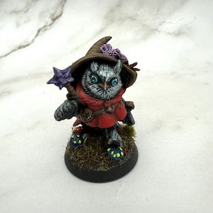 May include: A small, painted owl figurine wearing a brown hat with purple and orange flowers. The owl is wearing a red robe and holding a purple and silver wand. The owl is standing on a base with green grass and black mushrooms.