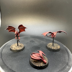 May include: Three miniature red dragon figurines with outstretched wings and detailed features. Each dragon is mounted on a small, round, textured base. The dragons are painted in shades of red, with visible wing details.