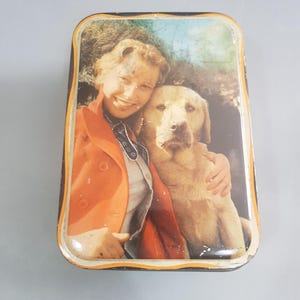 Può includere: Scatola di latta vintage con un'immagine colorata di una donna e un cane. La donna indossa una giacca rossa e il cane è un golden retriever.