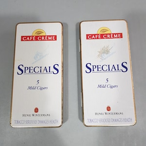 Op de afbeelding: Twee rechthoekige witgouden sigarettendoosjes. De dozen hebben de tekst "CAFÉ CRÈME" in rood, "SPECIALS" in blauw en "5 Mild Cigars". De onderkant van de dozen luidt "HENRI WINTERMANS" en "TOBACCO SERIOUSLY DAMAGES HEALTH".