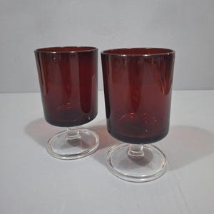 Könnte beinhalten: Zwei passende Trinkgläser mit tiefburgunderfarbenen Kelchen in zylindrischer Form. Jedes Glas wird von einem kurzen, klaren Glasstiel und einem kreisförmigen, flachen, klaren Glasfuß getragen, was dem Gefäß eine stabile Grundlage verleiht.