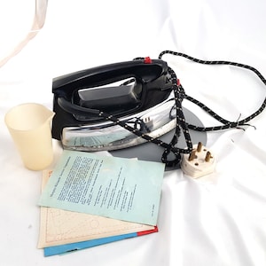May include: A black and silver electric iron with a black cord and a white plug. The iron is sitting on a white surface with a white cup and a sheet of paper with instructions.