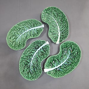 May include: Four ceramic dishes shaped like green cabbage leaves. Each dish features a textured, bumpy surface mimicking the veins and crinkles of a real leaf, with a prominent white central stem running through the middle of each individual piece.