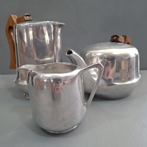 May include: A collection of vintage silver-toned serving items. The set comprises a creamer, a lidded sugar bowl, and a pitcher with a wooden handle. The items are displayed on a grey surface.