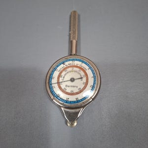 May include: A vintage map measuring tool with a circular face, a silver metal casing, and a knurled handle. The dial features white, blue, and orange sections with text reading Inches to Miles or Verstes, Centimeters to Kilometers, and Germany.
