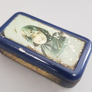 Könnte beinhalten: Eine blaue Metalldose mit einem verblassten Bild einer Frau, die einen Kapuzenumhang trägt. Der Text "REGISTERED TRADE MARK" ist auf der Dose sichtbar.
